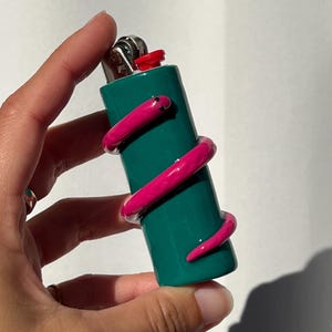 May include: A dark teal lighter featuring a three-dimensional, glossy pink snake wrapped around the cylindrical body. The snake has small black eyes and a smooth texture. The lighter has a metallic silver top mechanism and a bright red button.