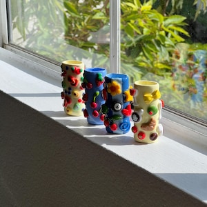 Pu&ograve; includere: Quattro vasi decorativi in ceramica, di forma cilindrica, con interni gialli. I vasi sono decorati con abbellimenti colorati, tra cui ciliegie e fiori. Sono esposti su un davanzale bianco, con uno sfondo naturale luminoso visibile attraverso la finestra.