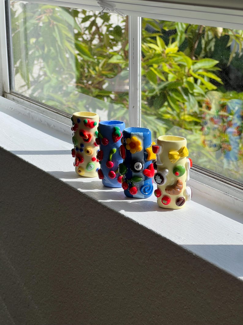 Pu&ograve; includere: Quattro vasi decorativi in ceramica, di forma cilindrica, con interni gialli. I vasi sono decorati con abbellimenti colorati, tra cui ciliegie e fiori. Sono esposti su un davanzale bianco, con uno sfondo naturale luminoso visibile attraverso la finestra.