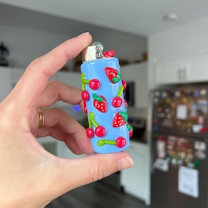 Pu&ograve; includere: Un accendino azzurro decorato con ciliegie e fragole rosse con steli verdi. L'accendino &egrave; tenuto in mano e lo sfondo &egrave; un interno sfocato.