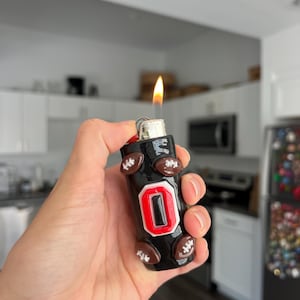 Pu&ograve; includere: Un accendino nero con un logo "O" rosso e bianco e dettagli a forma di pallone da football. L'accendino &egrave; acceso, mostrando una fiamma. L'accendino &egrave; tenuto in mano.