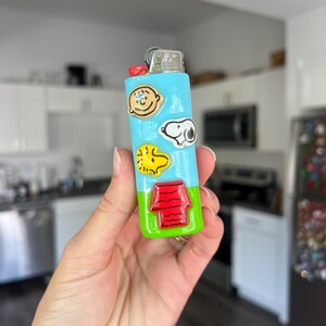 Pu&ograve; includere: Un accendino dipinto su misura con disegni di personaggi dei cartoni animati. L'accendino ha un corpo azzurro con immagini di Charlie Brown, Snoopy e Woodstock. Il fondo &egrave; verde con una cuccia rossa. La parte superiore &egrave; rossa.