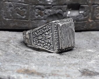 May include: A silver ring with a square, textured top and intricate, detailed designs on the band.