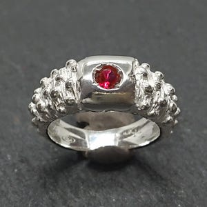 May include: A silver ring with a textured band and a square setting. The setting holds a small, round, red gemstone. The ring is displayed against a dark background.