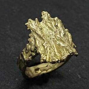 May include: A gold ring with a textured, organic design resembling a tree branch or a piece of rock. The ring is made of solid metal and has a unique, handcrafted look.