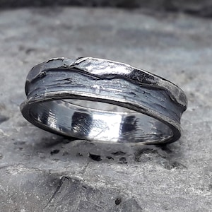 Puede incluir: Un anillo de banda de plata con un acabado texturizado y martillado. El anillo tiene una banda ancha con un borde ligeramente elevado.