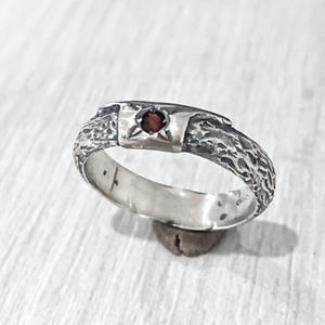 May include: A silver ring with a textured band and a square setting. The setting holds a small, round, red gemstone. The ring is displayed on a light-coloured surface.