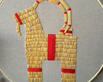 May include: An embroidered goat made of yellow yarn with a red ribbon around its neck, on a beige fabric background.