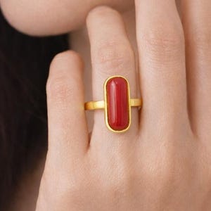 May include: A gold ring featuring a rectangular, vibrant red gemstone. The ring has a simple, elegant design with a polished gold band and a bezel setting that holds the red stone securely. The ring is worn on a finger.