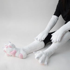 Op de afbeelding: Witte pluche kattenpoot handschoenen en beenwarmers met roze pootkussentjes. De handschoenen en beenwarmers zijn gemaakt van een zacht, pluizig materiaal. De poten hebben een realistisch ontwerp.