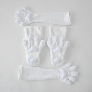 Può includere: Guanti e scaldabraccia bianchi in peluche a forma di zampa di gatto. I guanti presentano un design a zampa con cuscinetti delle dita arrotondati e un cuscinetto più grande sul palmo. Gli scaldabraccia sono lunghi e tubolari.