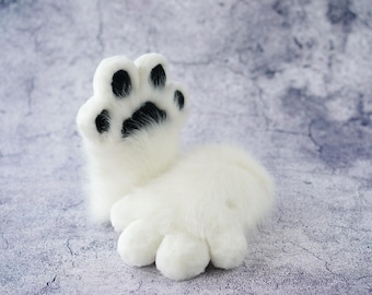 May include: A white plush toy paw with black paw pads. The paw is raised up and the other toes are curled under.