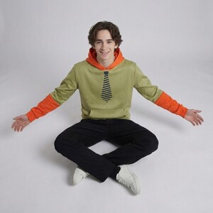 Pu&ograve; includere: Una felpa con cappuccio verde con cappuccio e maniche arancioni, con un design a cravatta nera. La persona indossa pantaloni neri e scarpe da ginnastica bianche. La felpa ha una tasca frontale.