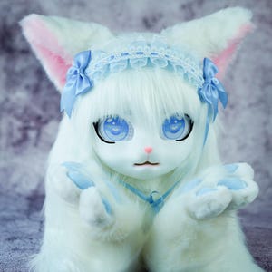 Op de afbeelding: Een wit, pluche speelgoeddier met blauwe ogen en een blauw en wit kanten hoofdband. Het speelgoed draagt een blauw lint om zijn nek en heeft zijn pootjes voor zich.