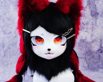 May include: A white, stylized mask with large, expressive eyes featuring heart-shaped pupils and orange irises. The mask has black hair, red and black fur accents, and silver hair clips. The overall design suggests a furry or cosplay aesthetic.