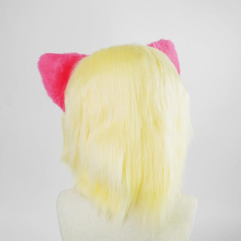 May include: A wig with straight, pale yellow synthetic hair featuring two bright pink, fuzzy, triangular cat ears attached to the top. The hair is shoulder-length and layered, providing a smooth texture throughout the style, set against a plain background.