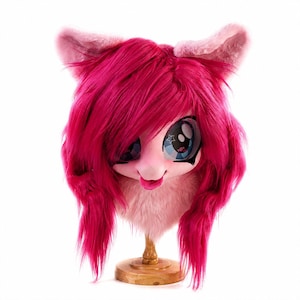 May include: A bright pink faux fur animal head mask featuring long, layered magenta hair, large expressive eyes with star accents, and pointed ears. The mask has a light pink muzzle and is displayed on a wooden stand against a white background.