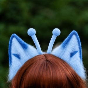 Puede incluir: Una diadema con dos orejas de piel sintética de color azul claro con detalles triangulares interiores en azul oscuro y dos tallos de antena delgados de color azul claro rematados con pequeñas esferas, sobre cabello castaño rojizo.