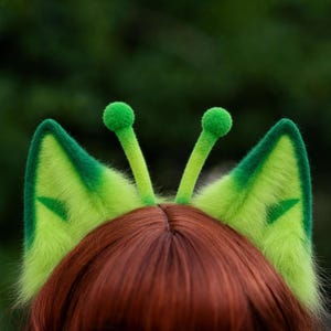 May include: A pair of bright lime green fuzzy cat ears with darker green trim and triangular accents. Two thin, upright antennae with small green pom-pom tips extend from the center between the ears, all set against a dark background.