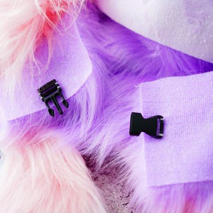 May include: Close-up of a fluffy, multi-coloured fabric with pink, purple, and white hues. Two black plastic buckles are attached to the fabric, one with a three-pronged design and the other with a single-prong design. The fabric appears soft and textured.