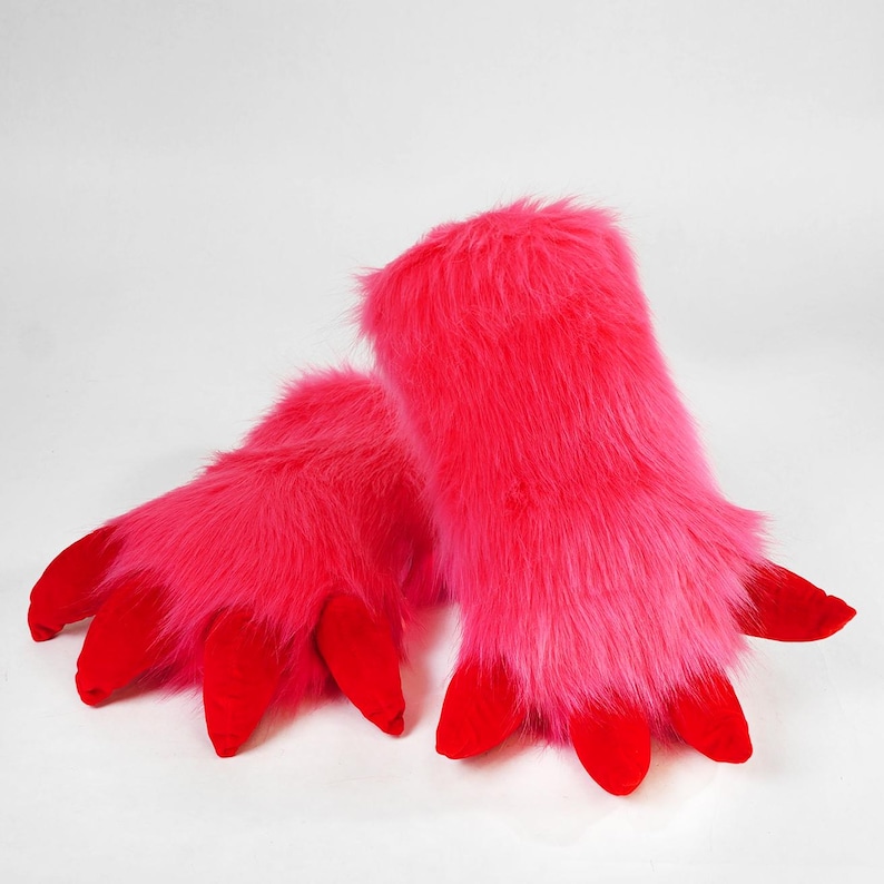 May include: A pair of bright pink faux fur gloves featuring long, shaggy synthetic fibers and distinct red fabric claws at the fingertips. The gloves have a thick, plush texture and a vibrant, saturated color palette, designed as hand coverings.