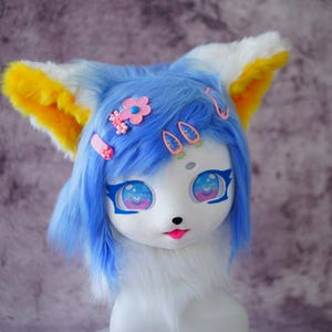 May include: A blue and white furry mask with large, expressive eyes and a pink tongue. The mask has blue hair, yellow and white ears, and is adorned with pink flower and barrette hair accessories. The overall design is whimsical and cartoonish.