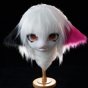 May include: A white and black furry mask with large, expressive eyes and a pink nose. The ears have black and pink accents. The mask is mounted on a wooden stand, showcasing its detailed design and craftsmanship. The overall aesthetic is reminiscent of anime or cosplay.