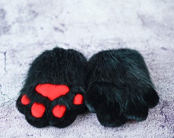 May include: A pair of black faux fur paw gloves with bright red paw pads. The gloves are designed to resemble animal paws, with a fluffy texture and rounded shape. The red pads are visible on the underside of the paws.