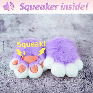 Pu&ograve; includere: Un paio di scaldamani in peluche viola a forma di zampa di gatto con cuscinetti bianchi. Gli scaldamani hanno uno squeaker all'interno e il testo "Squeak!" &egrave; scritto su uno degli scaldamani.