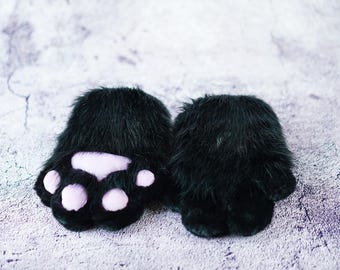 May include: A pair of black, furry paw-shaped mittens with light pink paw pads. The mittens are designed to resemble animal paws, with a soft, plush texture. The background is a mottled gray and white.
