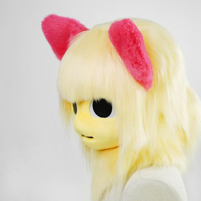 May include: A full-head mask featuring pale yellow synthetic hair with bangs and two bright pink fuzzy ears on top. The mask has a smooth yellow face with large, circular black eye openings and a small mouth opening, designed for full coverage.