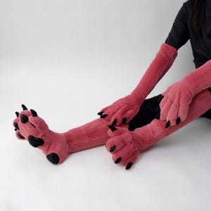 Può includere: Guanti e piedi rosa con artigli di mostro in peluche con artigli neri. I guanti si estendono sulle braccia e i piedi sono progettati per essere indossati come pantofole. Il set è realizzato in materiale morbido e soffice.