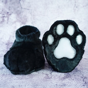 May include: A pair of black furry slippers with paw-shaped toes. The slippers have a white paw print design on the top.
