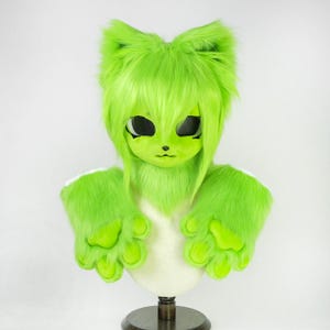 May include: A bright lime green furry animal head mask featuring large pointed ears, long shaggy hair, and black eye sockets. The set includes two matching lime green furry paw gloves with prominent padded fingers, displayed on a mannequin stand.