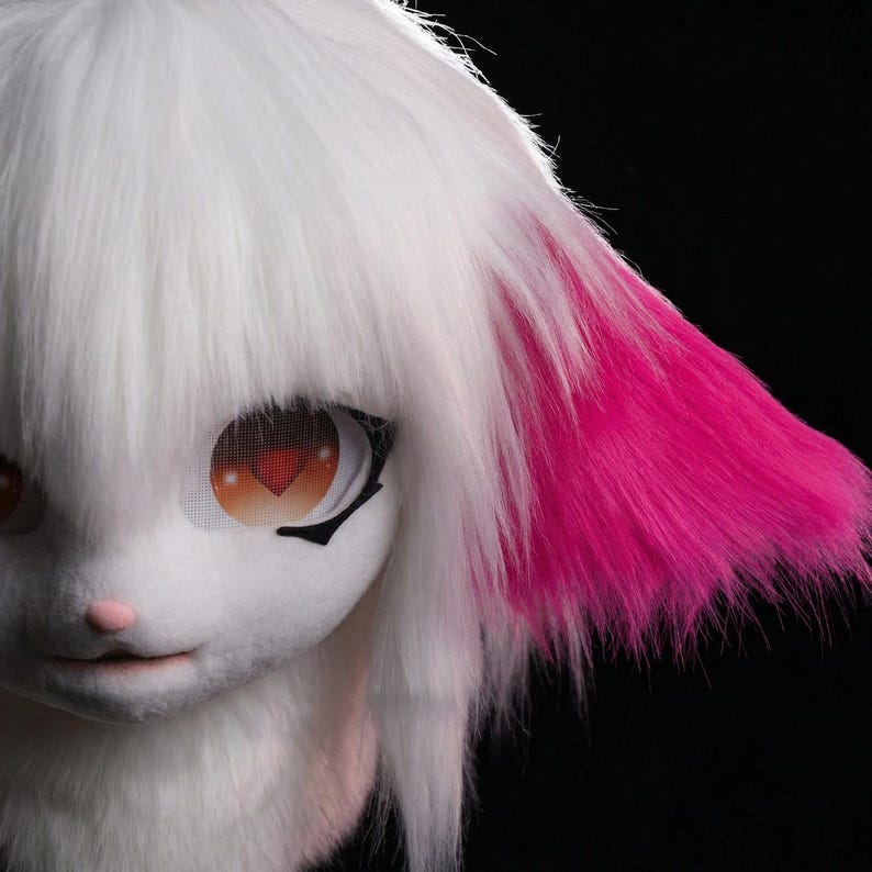 May include: A white furry mask with long, white hair and bright pink accents on the ear. The mask features large, expressive eyes with a heart-shaped pupil and a small pink nose. The mask is designed to resemble a cartoon character.