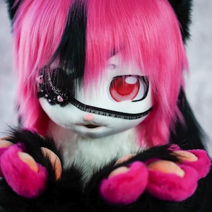 May include: A pink and black furry character with a white face and red eye. The character is wearing a black eye patch with silver and black embellishments. The character has pink paws with black claws.