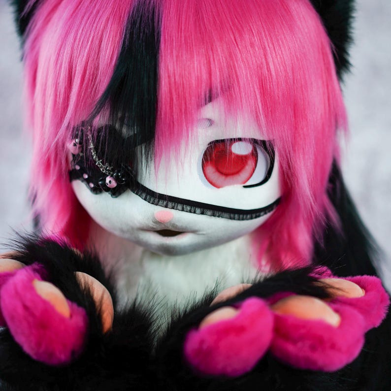 May include: A pink and black furry character with a white face and red eye. The character is wearing a black eye patch with silver and black embellishments. The character has pink paws with black claws.