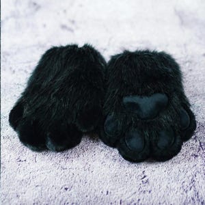May include: A pair of black faux fur paw gloves. The gloves have a plush, furry texture with a smooth, black paw pad and individual toe details. The gloves are designed to resemble animal paws.