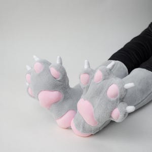 May include: Gray plush paw slippers with pink paw pads and white claws. The slippers are designed to resemble a cat's paws. The slippers are a fun and cozy accessory.