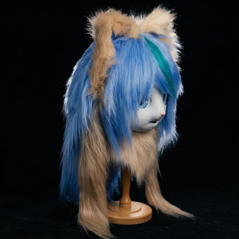 May include: An anime-style mask with blue and brown fur. The mask features large blue eyes, a small pink nose, and a light-colored face. The mask is mounted on a wooden stand against a black backdrop.