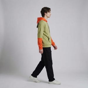 Pu&ograve; includere: Una felpa con cappuccio verde chiaro con maniche e cappuccio arancioni, abbinata a pantaloni neri e scarpe da ginnastica bianche. La felpa ha una tasca frontale e un design nero sul lato. La persona cammina in uno studio.