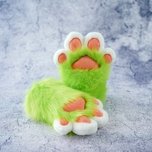May include: A pair of plush green paw gloves with white and peach-colored accents. The gloves have a furry texture and are designed to resemble animal paws. The gloves are set against a light gray background.