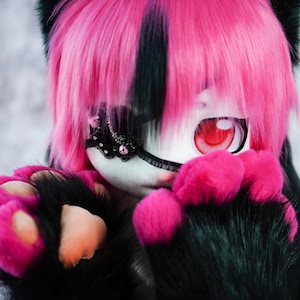 May include: A plush toy with pink and black fur, a black eye patch with pink and white accents, and one red eye. The toy has pink paws and is wearing a black collar.