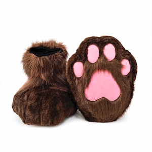 May include: A pair of brown faux fur slippers shaped like animal paws. The slippers feature long, shaggy brown synthetic fur and large, smooth, light pink fabric pads on the soles, representing the paw pads and toes of an animal.