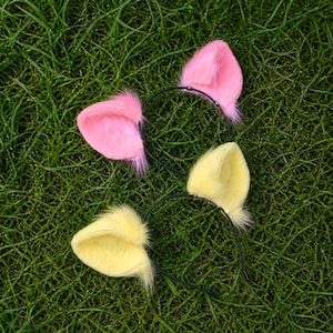 May include: Two thin black metal headbands, each featuring a pair of plush, fuzzy animal ears. One headband has bright pink ears, while the other headband has soft yellow ears. The ears are constructed with a textured, faux fur material.