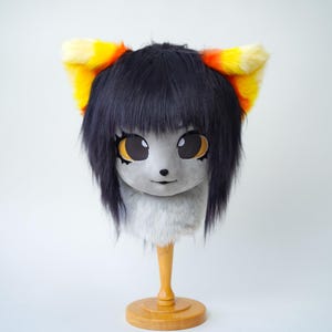 Könnte beinhalten: Eine pelzige, grau-weiße Katzenmaske mit großen, ausdrucksstarken Augen und schwarzen Haaren. Die Ohren sind gelb und orange. Die Maske wird auf einem Holzständer vor weißem Hintergrund präsentiert. Für Cosplay oder Kostüme.