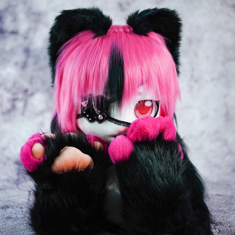 May include: A plush toy with black fur, pink paws, and a pink and black wig. The toy has a white face with a black eye patch and one red eye.