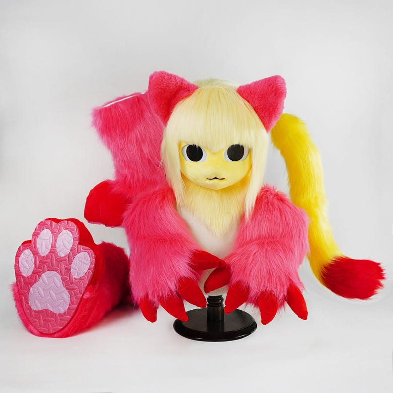 May include: A plush animal head mask with blonde hair, pink ears, and large black eyes, accompanied by matching pink furry hand paws with red claws and a pink paw pad, plus a long yellow and red tail, all displayed on a black stand.