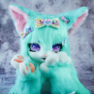 May include: A turquoise blue furry plush toy with pink ears and a yellow, blue and white bow. The toy has large purple eyes and a pink nose. The toy is sitting on a grey textured background.