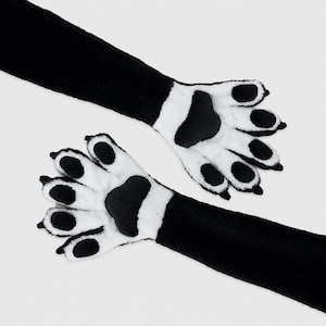 May include: A pair of long, plush gloves featuring a black forearm section and white hand section with black paw pads and black claw-like tips. The design mimics animal paws with soft, fuzzy fabric covering the entire length of the gloves.